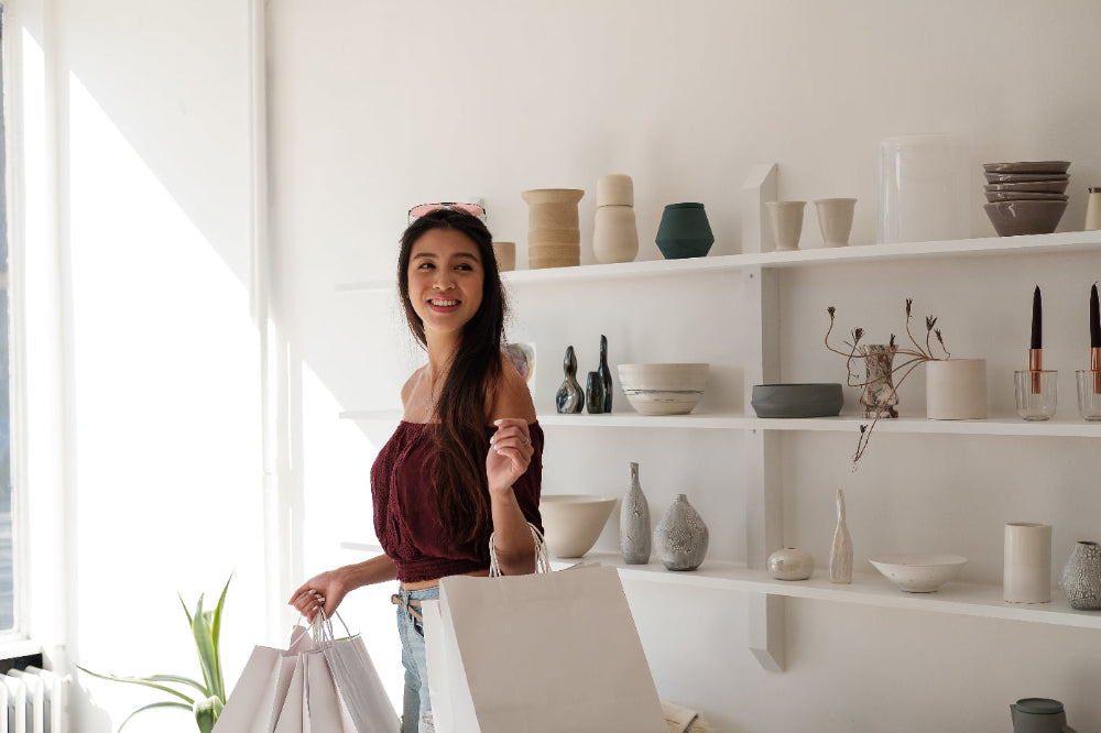 Woman holding shopping bags in a room with shelves displaying various shopping items.